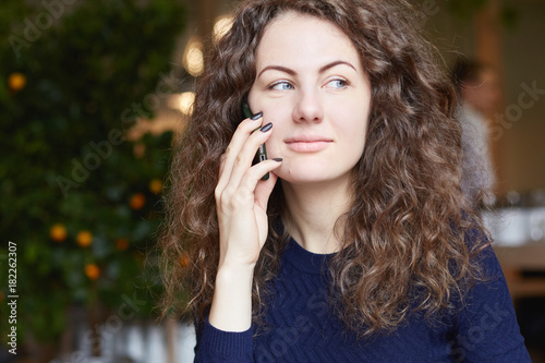 Beautiful student lady with charming smile wearing blue sweater talking on mobile phone while studying at cafe, using wireless internet connection on laptop computer, waiters moving in background.