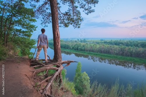 Landscape at dusk on the high bank of the river with a guy standing on the roots of a large pine