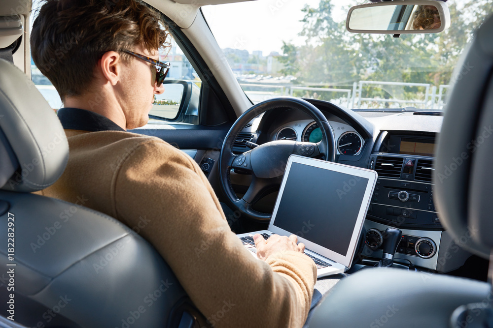 Back view portrait of successful modern entrepreneur wearing sunglasses working, using laptop sitting in luxury car, copy space