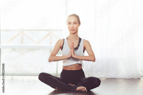Portrait of beautiful young woman working out against window, doing yoga and pilates exercises, meditating and sitting in baddha konasana, butterfly pose. Full length shot