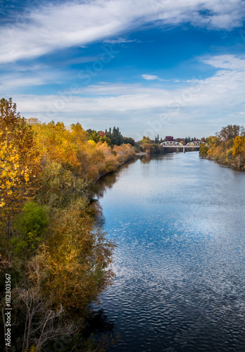 River view with trees in autumn colors on both banks. A bridge is crossing the river in the distance. A blue sky with clouds is in the background. The water is reflecting the blue sky and white clouds