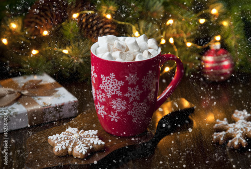  Close-up of a mug with a hot drink, marshmallow and gingerbread. Christmas decor with a bright garland and fir tree. Selective focus, shallow depth of field, background
