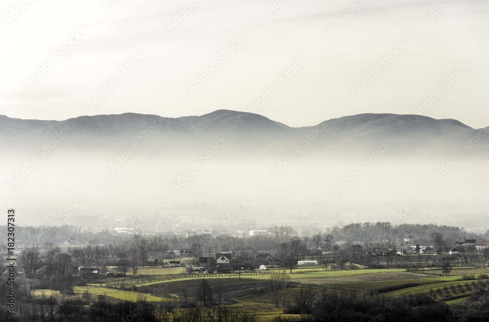 Fototapeta premium foggy rural landscape of village with green fields and mountains hill top