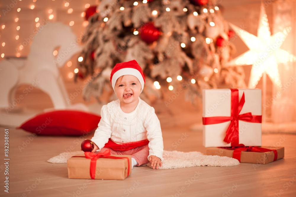 Laughing baby girl 1 year old having fun with Christmas presents over Christmas lights in room. Looking at camera. Childhood. Stock Photo Adobe Stock