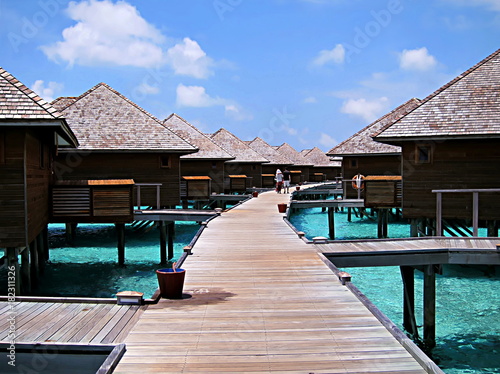 Fototapeta Naklejka Na Ścianę i Meble -  Walkway with water bungalows on both sides and with a couple of tourists strolling around, in Maldives, on a sunny day