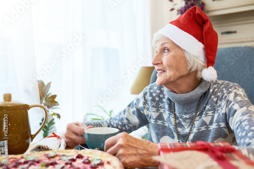Portrait of smiling senior woman wearing Santa hat looking to window pensively enjoying  Christmas dinnerat home  sitting at table and drinking tea, copy space