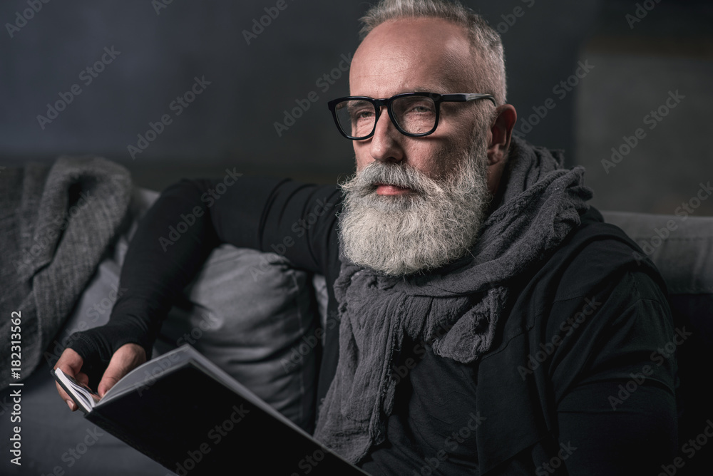 Portrait of thoughtful bearded grandfather reading interesting book while sitting on cozy sofa. Rest concept Portrait of thoughtful bearded grandfather reading interesting book while sitting on cozy sofa. Rest concept