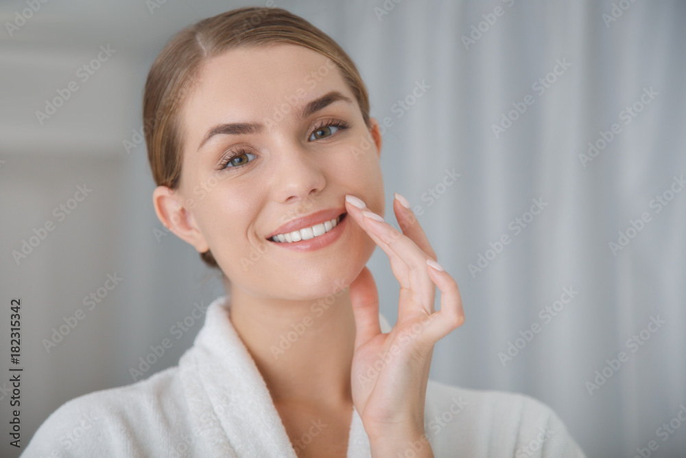 I like my skin. Portrait of happy young woman is touching her face while standing in spa bathrobe. She is looking at camera and smiling I like my skin. Portrait of happy young woman is touching her face while standing in spa bathrobe. She is looking at camera and smiling
