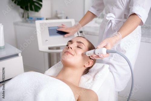 Side view of happy young woman getting cavitation rejuvenating skin treatment at spa. She is lying on massage table and smiling. Beautician is touching monitor screen while holding tool near female