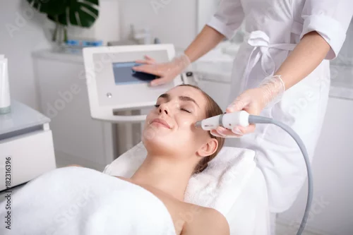 Obraz Side view of happy young woman getting cavitation rejuvenating skin treatment at spa. She is lying on massage table and smiling. Beautician is touching monitor screen while holding tool near female