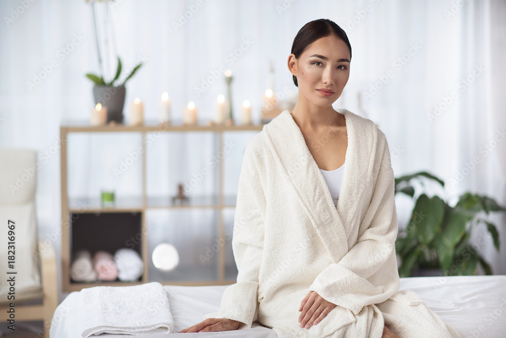 Have rest. Portrait of elegant relaxed young asian woman in white bathrobe is sitting on massage table at spa salon with candles in background. She is looking at camera thoughtfully. Copy space Have rest. Portrait of elegant relaxed young asian woman in white bathrobe is sitting on massage table at spa salon with candles in background. She is looking at camera thoughtfully. Copy space