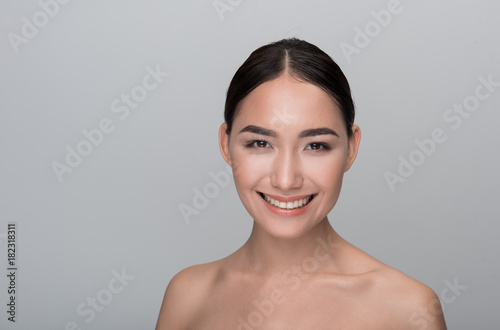Daily skincare. Portrait of optimistic young naked asian girl with fresh and clean face. She is looking at camera with wide smile. Isolated background and copy space in the left side
