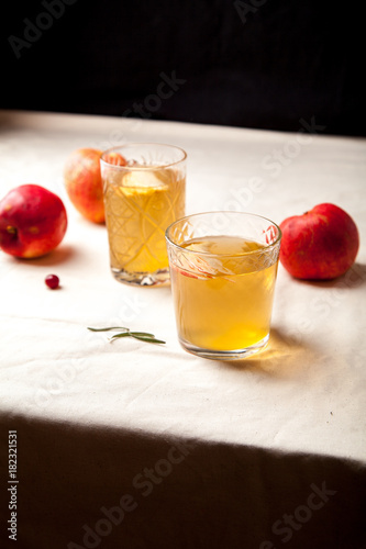 Two vintage glasses with apple cider on black background. Christmas beverages concept. Two red apples and rosemary sprig aside.  Warm backlight. Vertical composition.