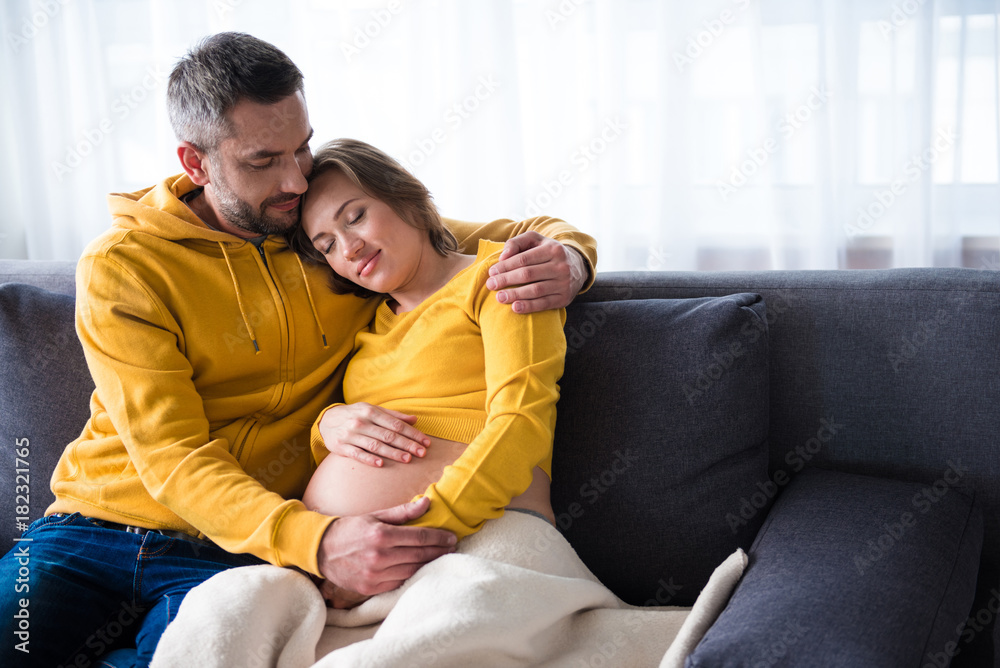 Feeling safe. Portrait of careful man embracing his expectant wife with love. Woman is leaning head on his shoulder while sitting on couch. Her eyes are closed with pleasure Feeling safe. Portrait of careful man embracing his expectant wife with love. Woman is leaning head on his shoulder while sitting on couch. Her eyes are closed with pleasure
