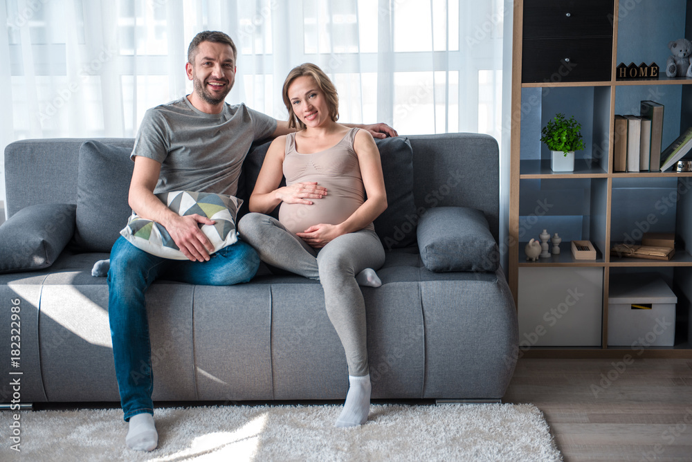 Happy family. Full length portrait of excited married couple sitting on sofa and smiling. Pregnant woman is touching her belly with joy