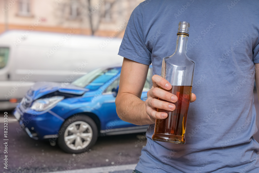 Traffic accident and alcohol concept. Drunk driver is holding bottle with alcohol in hand. Crashed car in background. Traffic accident and alcohol concept. Drunk driver is holding bottle with alcohol in hand. Crashed car in background.
