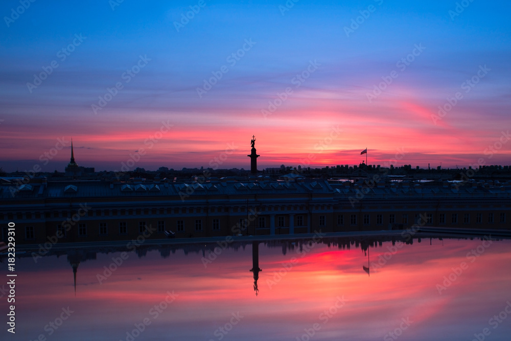 reflection of the Alexander Column Palace Square on the glass against the background of a red and blue evening sky /A view of the Alexander Column and the setting sun from the window of the building 