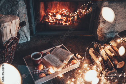 Warm cozy fireplace with real wood burning in it. Magical atmosphere. Cup of hot drink and book ready for evening relax. Cozy winter concept. Christmas and travel background with space for your text.