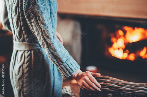 Woman in warm knitted cardigan sweater by fireplace. Woman relaxes by warm fire with a cup of hot drink and warming up her feet in woollen socks. Cozy atmosphere. Winter and Christmas holidays concept