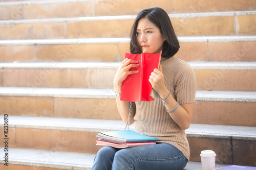 Portrait of Attractive Asian Student Woman sitting at outdoor place. Woman holding Book with Serious Emotion. People with Education Concept.