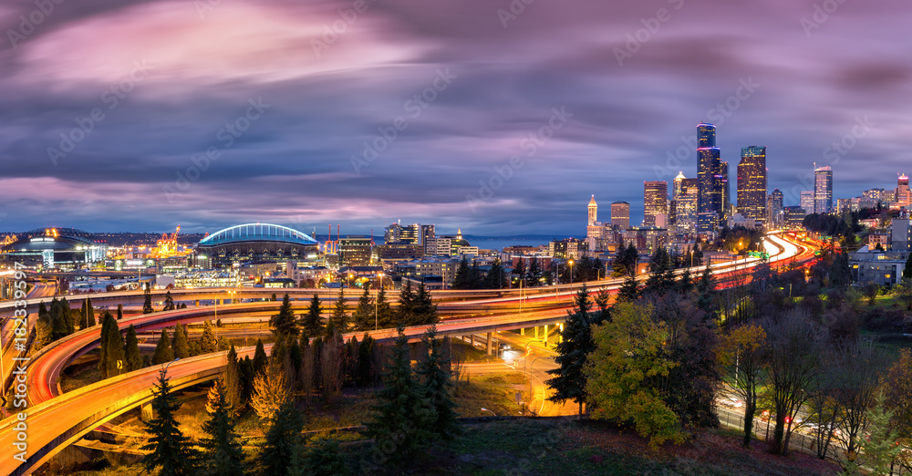 Seattle cityscape at dusk with skyscrapers, winding highways parks and sports arenas under a dramatic sky.