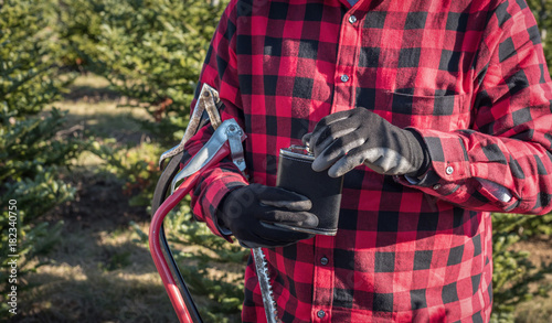 man in red plaid shirt holding saw and flask at christmas tree farm