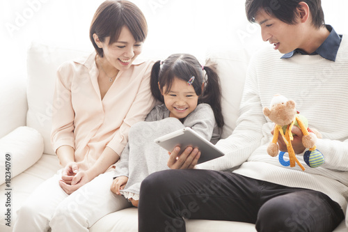 Parents and daughter are sitting on the sofa and using a digital tablet together.