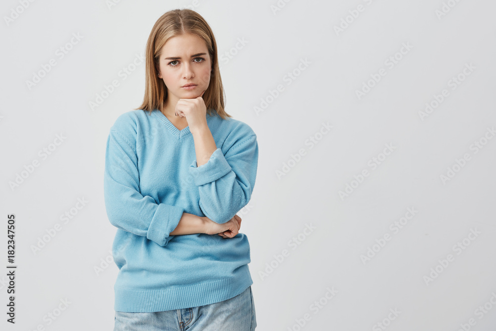 Beautiful caucasian dissatisfied female with straight fair hair wearing casual clothes looking at camera with anger, holding her hands under her chin, dissatisfied with actions of her boyfriend. Beautiful caucasian dissatisfied female with straight fair hair wearing casual clothes looking at camera with anger, holding her hands under her chin, dissatisfied with actions of her boyfriend.