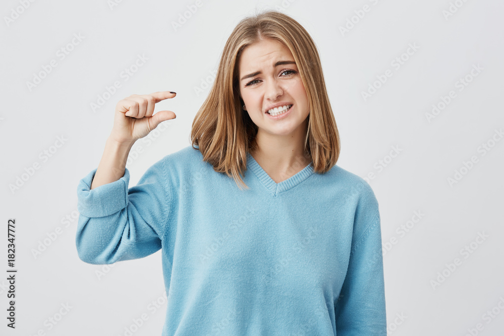 Fototapeta premium European young female wearing blue sweater with blonde hair showing something small in size with hands, gesturing, frowning brows and showing her teeth. Blonde demonstrating size of something.