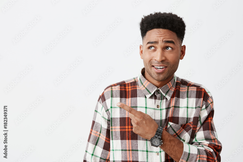 Close up of young handsome dark-skinned american male student with curly hairstyle in checkered long sleeve shirt pointing aside with index finger on white wall with cynical face expression Close up of young handsome dark-skinned american male student with curly hairstyle in checkered long sleeve shirt pointing aside with index finger on white wall with cynical face expression