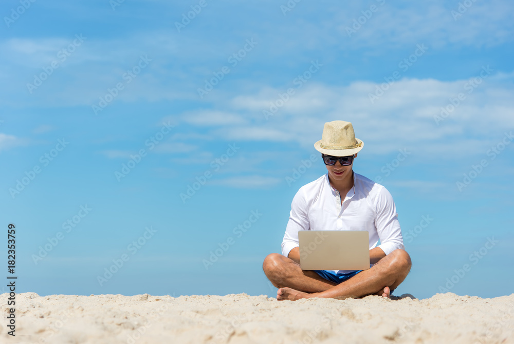 Lifestyle young asian man working on laptop while sitting on the beautiful beach, freelance working on holiday summer, blue sky background.  Summer Concept