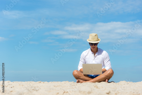 Lifestyle young asian man working on laptop while sitting on the beautiful beach, freelance working on holiday summer, blue sky background. Summer Concept