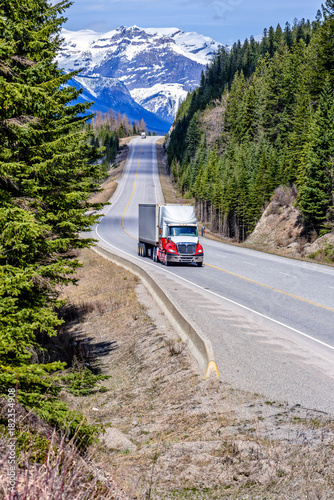 Trucks drive along an asphalt road among the forests and snow-capped mountains