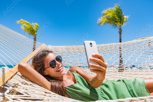 Summer beach lifestyle young woman using phone app texting on smartphone relaxing at tropical Caribbean resort on hammock. Mobile wifi internet on travel holidays. Vacation fun.