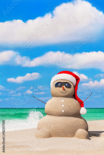 Sandy Snowman in Christmas Santa hat and sunglasses at tropical beach against waves splashes