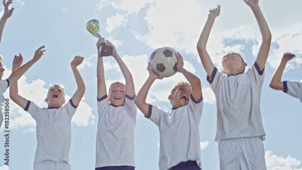 Low angle of ecstatic boys from junior soccer league holding gold cup and ball and jumping up and down in celebration after winning match