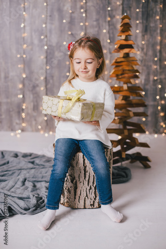 Happy little girl in a white sweater and blue jeans posing near christmas tree © polinaloves