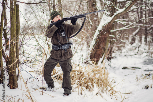 Male hunter in camouflage, armed with a rifle, standing in a snowy winter forest with duck prey