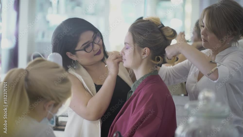 For young woman doing hairdo, makeup and manicure in beauty salon. Artist applies purple shadows on eyelids, stylist makes curls with help of curling iron and master in mask does manicure. Three