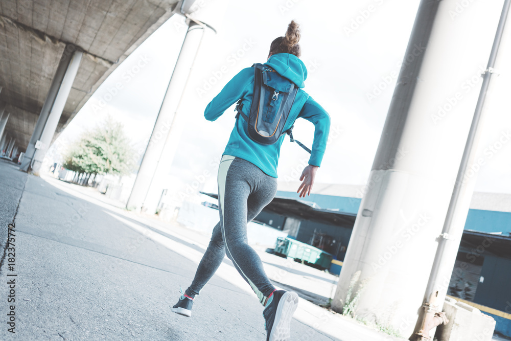 Athlete beautiful girl with waterbackpack runnig in the morning on the city street. Sport tight