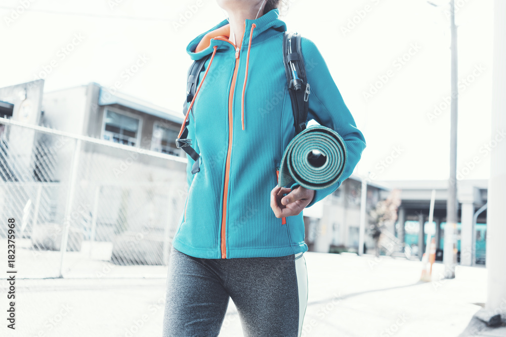 Obraz premium Athlete beautiful girl with backpack, headphones, yoga mat going to sport club on the city street. Sport tight clothes. Blurry background.