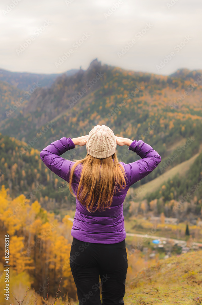 A girl in a lilac jacket looks out into the distance on a mountain, a view of the mountains and an autumnal forest by an overcast day, free space for text.
