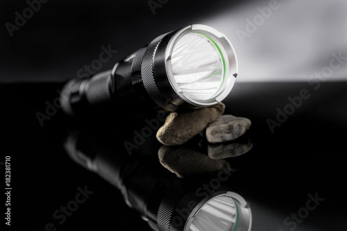 Black flashlight with lights open on stones mirroring on black background.