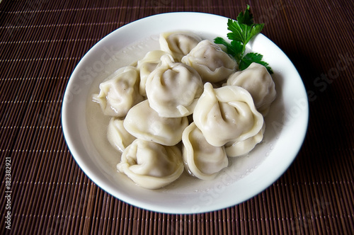 Home / purchased dumplings in a broth with greens. Dumplings are laid out in a deep white bowl. The bowl is on the stand. Day light. Dumplings are decorated with parsley leaves.