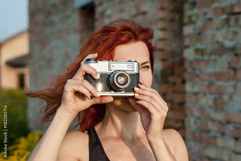 Red-haired girl makes a photo vintage film camera on the background of a brick wall