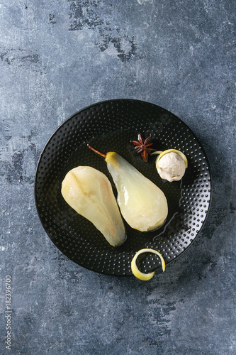 Traditional dessert sliced poached pears in white wine served in black plate with syrup, ice cream and lemon zest over blue texture background. Top view with space