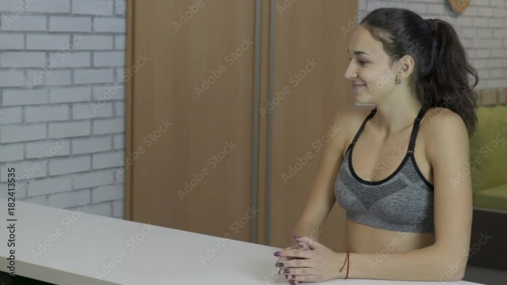 Young female at reception desk in sports club. Woman approaches, gives number to administrator with friendly smile and receives soft towels. Fitness lady with long tail dressed in stylish top with Young female at reception desk in sports club. Woman approaches, gives number to administrator with friendly smile and receives soft towels. Fitness lady with long tail dressed in stylish top with