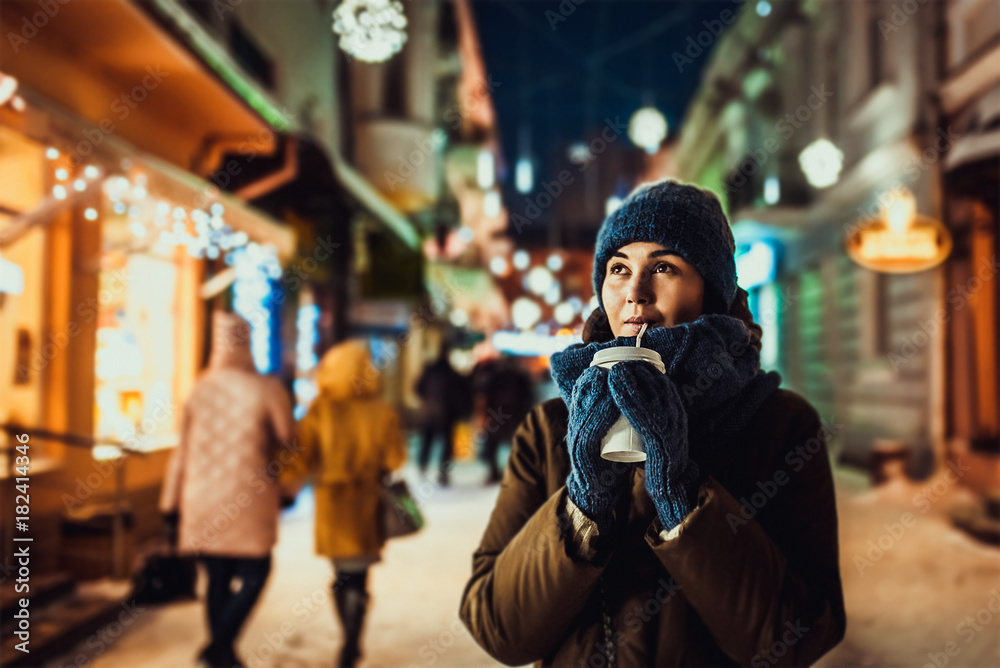 Pretty girl with coffee outdoor in cold weather with christmas lights behind