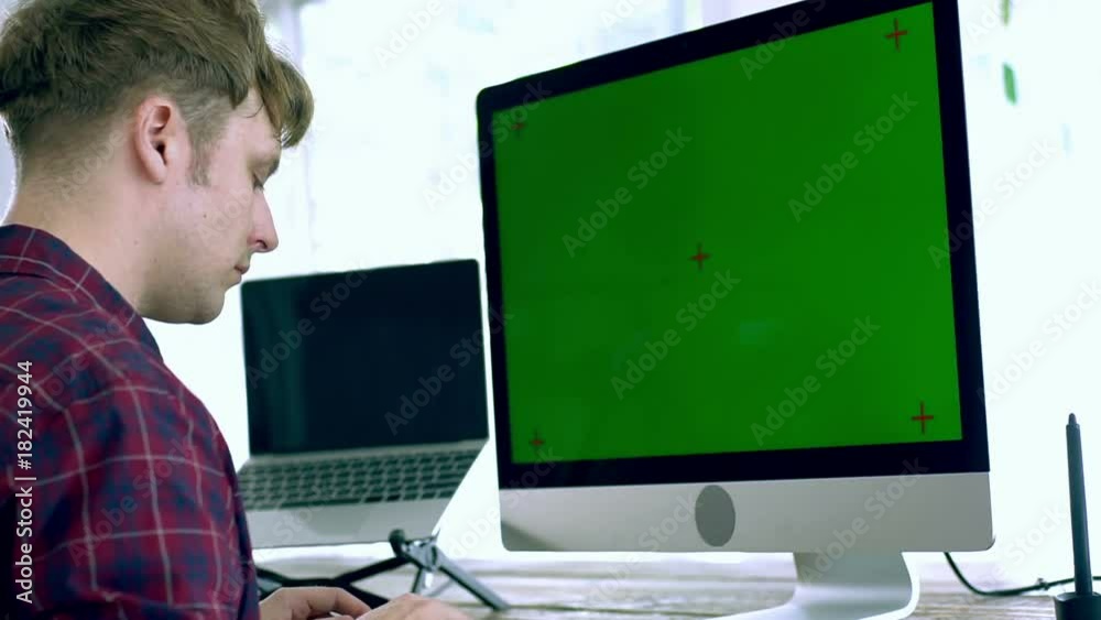 A caucasian male freelance designer, programer working at Home with all in one unit computer with green screen, laptop, a pen tablet and other electronics peripheral on the wooden table on a bright mo A caucasian male freelance designer, programer working at Home with all in one unit computer with green screen, laptop, a pen tablet and other electronics peripheral on the wooden table on a bright mo
