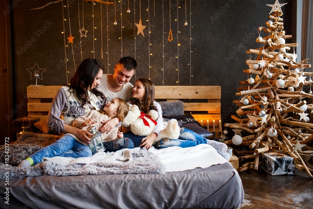 A family of four hugging in front of the christmas tree on the bed with candles and gifts. New year's eve. Cozy holiday at the fir-tree. love, happiness and big family concept